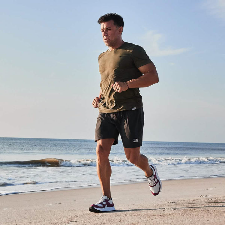 A man jogs on a sandy beach by the ocean under a clear blue sky, capturing the essence of a Men's Rough Runner - Baton Rouge. He wears a green T-shirt, black shorts, and sneakers from GORUCK. The waves gently lap at the shore in the background.