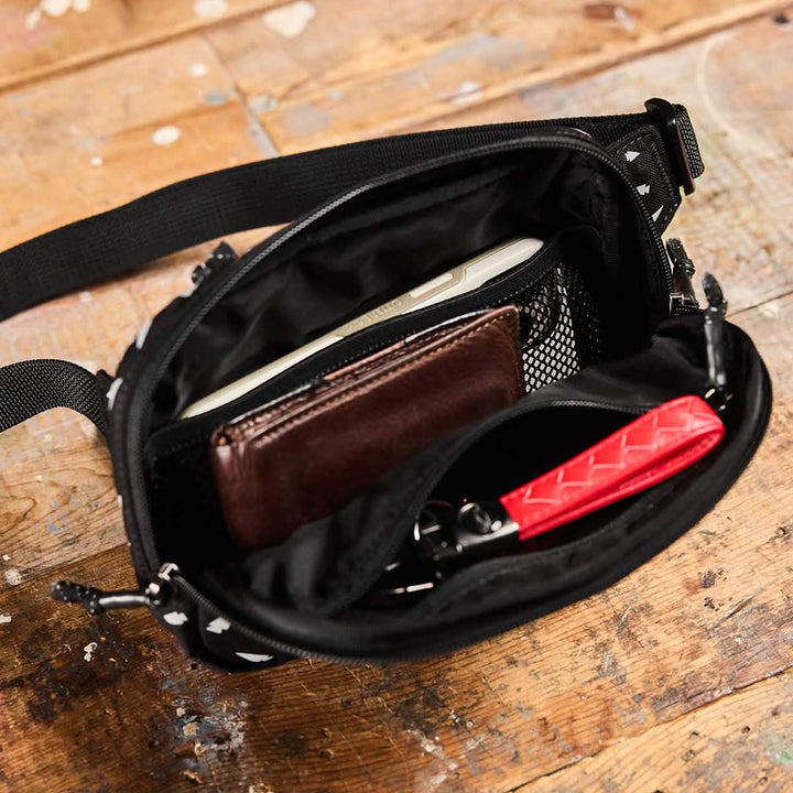 A small black GORUCK Belt Bag sits on a wooden surface, its contents neatly organized: a smartphone, brown wallet, and a red item with a grid pattern. The compact gear bag features adjustable webbing and multiple compartments, including a zippered section with mesh pockets.