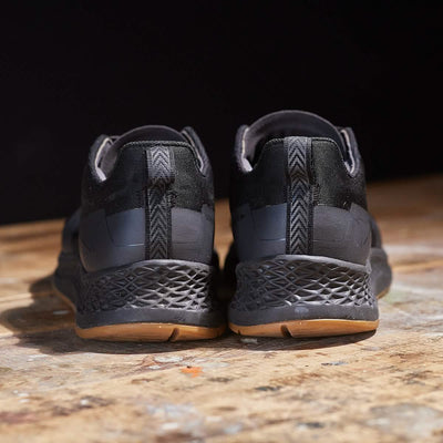 A pair of Women's Rough Runner - Black + Gum sneakers by GORUCK, featuring textured soles and brown accents, is showcased from the back. With an EVA midsole for enhanced comfort, these sneakers rest elegantly on a wooden surface against a dark background.