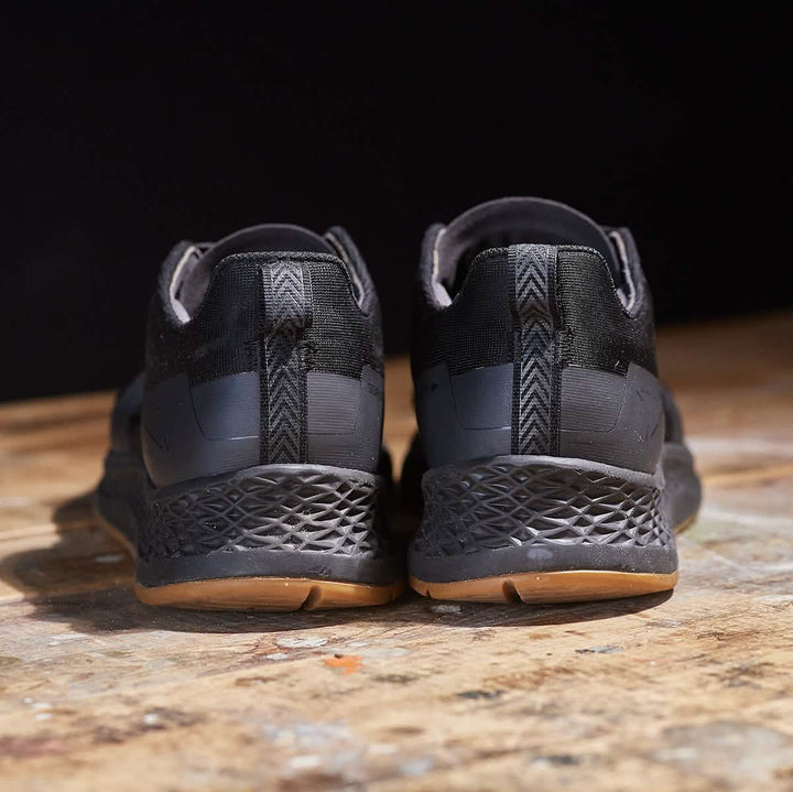 A pair of Women's Rough Runner - Black + Gum sneakers by GORUCK, featuring textured soles and brown accents, is showcased from the back. With an EVA midsole for enhanced comfort, these sneakers rest elegantly on a wooden surface against a dark background.