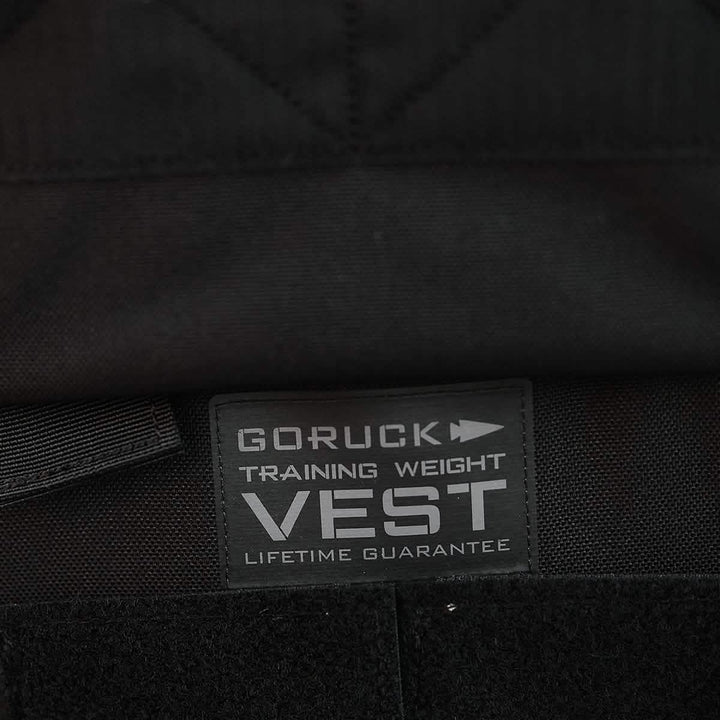 A close-up of a black fabric tag on a garment features the text "Training Weight Vest 2.0 LIFETIME GUARANTEE" above GORUCK's signature arrow logo, ideal for individuals looking for an adjustable weight vest experience.