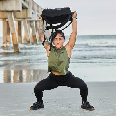 A person exercises on the beach, performing an overhead squat with the Ruck Clubs Starter Kit - Heavy tucked into their backpack. They wear a green sleeveless shirt and black leggings as the ocean and a pier form the scenic backdrop, training for an upcoming GORUCK event.