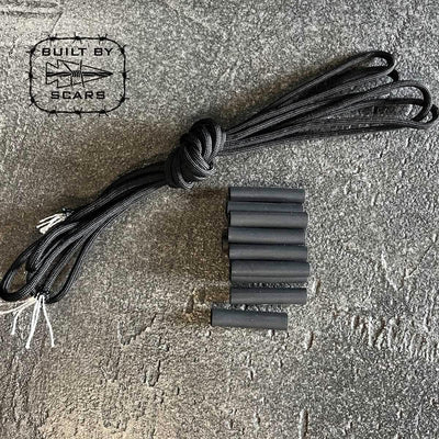 On a textured surface, black zipper pulls from the Paracord Kit - Built By SCARS by GORUCK are artfully arranged, featuring knots and several heat shrink tubes for an ideal DIY ruck upgrade. The iconic Built by SCARS logo in the upper left corner signifies rugged craftsmanship.
