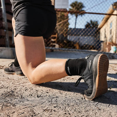 A person kneels on one knee outdoors, highlighting the Women's Ballistic Trainers in Black + Gum with a Black Reflective Spearhead by GORUCK. The scene includes a chain-link fence and a building framed by palm trees, with sunlight casting shadows on the concrete ground, making these trainers ideal for functional fitness.