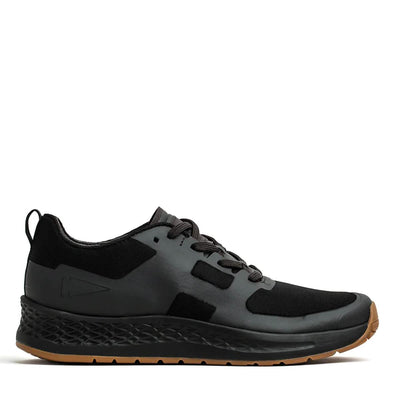 The Men's Rough Runner - Black + Gum by GORUCK is a black athletic shoe with a sleek design, characterized by a textured sole and subtle overlays. It features black laces and displays a small logo on the side. The shoe includes a Gradient Density EVA Midsole for enhanced comfort, set against a striking white background.