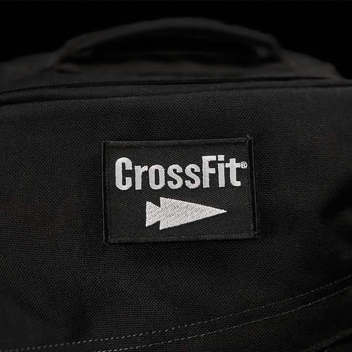 A close-up view of the Patch - GORUCK x CrossFit from GORUCK-DEMO showcases the word 'CrossFit' and a stylized arrow logo in white. The patch is stitched onto a black fabric surface, likely part of a bag or clothing item, with a dark and out-of-focus background.