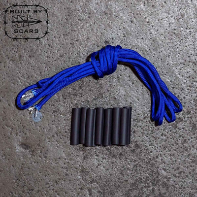 Neatly coiled next to a row of cylindrical black objects on a textured concrete surface is a paracord rope. In the upper left corner, an emblem with "Built By SCARS" is visible, highlighting the ideal setting for DIY ruck upgrades or crafting with the Paracord Kit by GORUCK.