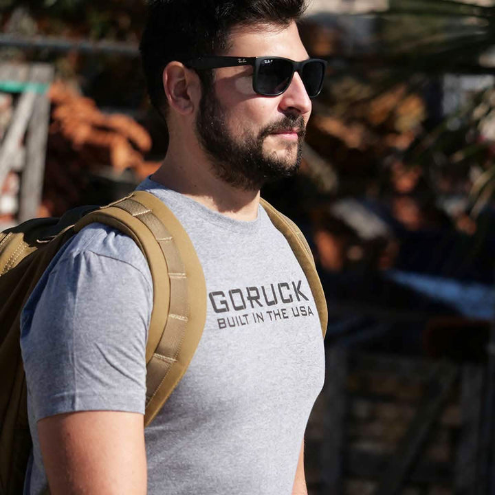 A man with a beard, wearing sunglasses and a gray Tri-Blend Reverse Flag Tee from vendor-unknown, strides confidently outdoors. His tan backpack complements the blurred backdrop of foliage and structures, embodying courage and respect.