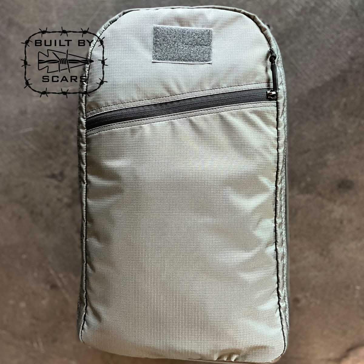 The Bullet 15L Robic Nylon backpack in gray, manufactured by GORUCK, is showcased against a textured background. It includes a front zippered pocket and prominently displays the "Built by SCARS" logo featuring barbed wire and a shark on the left side.