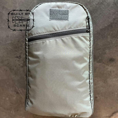 The Bullet 15L Robic Nylon backpack in gray, manufactured by GORUCK, is showcased against a textured background. It includes a front zippered pocket and prominently displays the "Built by SCARS" logo featuring barbed wire and a shark on the left side.