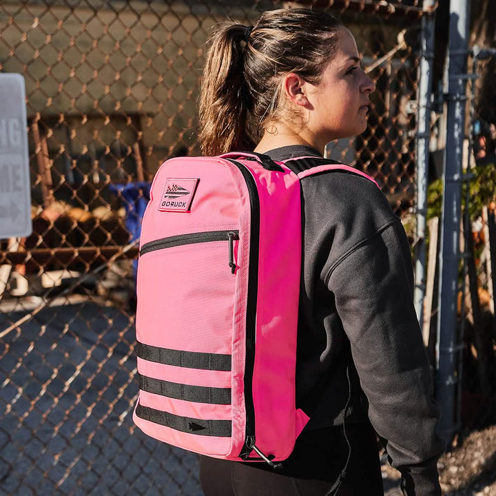 A woman in a black sweatshirt stands outdoors near a chain-link fence, wearing the Bullet Ruck - Classic - Hot Pink daypack.