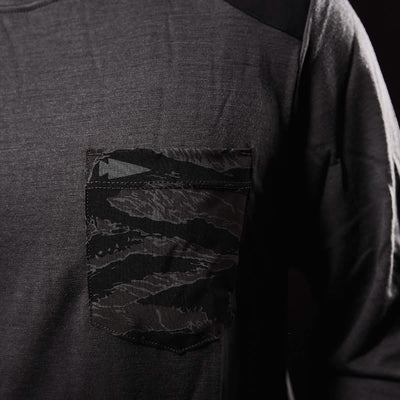 Close-up of the Men’s Commando Long Sleeve - Merino Wool by GORUCK, featuring a dark gray hue with a single front pocket. The pocket showcases a black and gray camo pattern, subtly contrasting with the plain fabric and evoking the rugged style reminiscent of a British Commando Sweater. Displayed against a dark background, the shirt's refined design stands out beautifully.