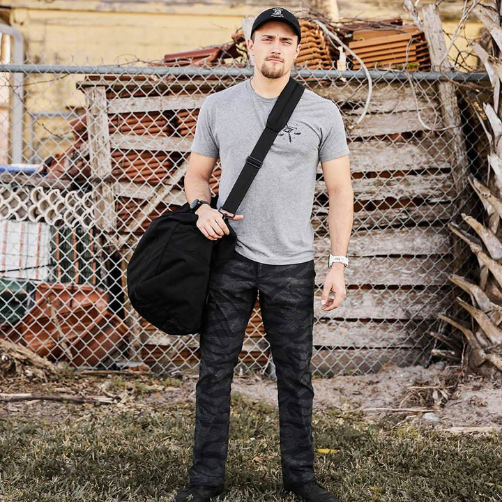Standing on the grass, a person is dressed in a gray t-shirt, camouflage pants, and a black cap. They are carrying the sleek Kit Bag with a shoulder strap from GORUCK. Behind them is a chain-link fence and stacked wooden pallets.