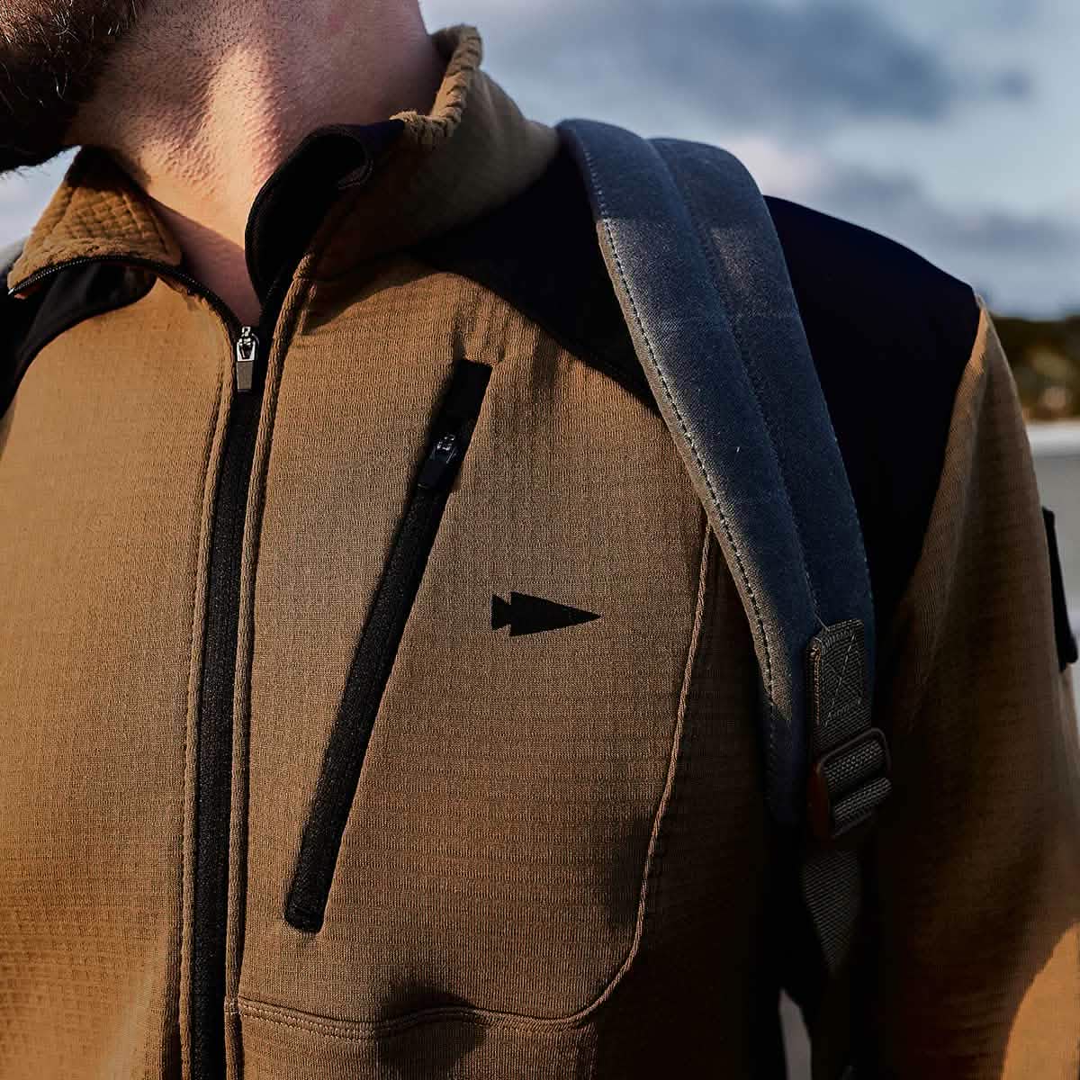 A close-up of a person wearing the GORUCK Men’s Half Zip jacket, featuring a brown design with black accents and a triangle logo, crafted from ToughDry Fabric, while carrying a gray strap backpack. The sky is partly visible in the background.