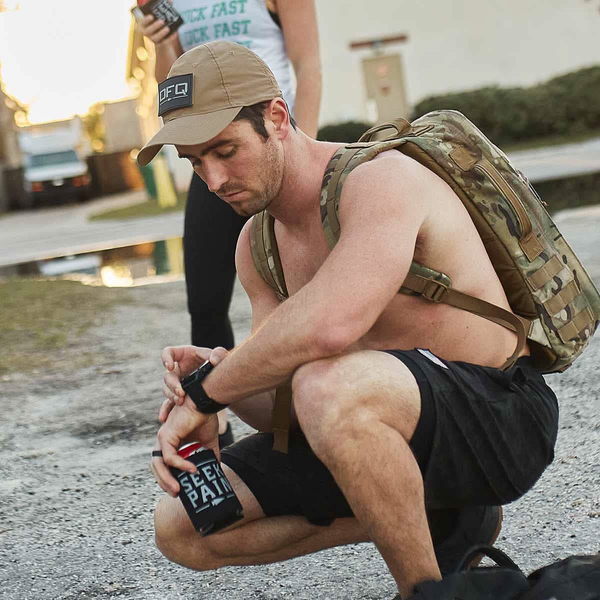 A shirtless man in a GORUCK Performance TAC Hat - TOUGHDRY and black shorts kneels on a gravel surface, wearing a camouflage backpack. He holds a black can labeled Seek Pain and checks his watch. A blurred building and truck are in the background, completing this rugged scene.