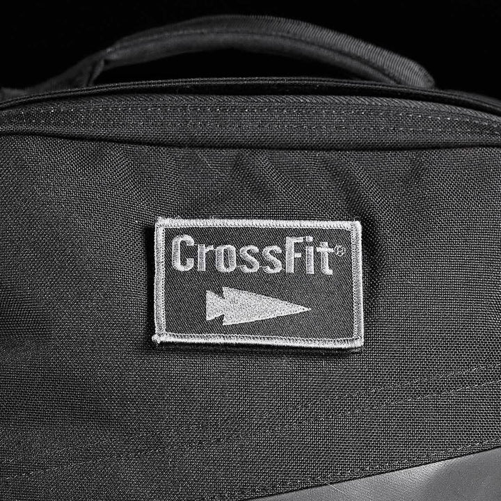 A black fabric bag prominently displays a rectangular patch embroidered with the CrossFit logo and an arrow symbol in white. The stitching accentuates the central patch, seamlessly merging durability with style, embodying the rugged design principles of GORUCK-DEMO's Patch - GORUCK x CrossFit.