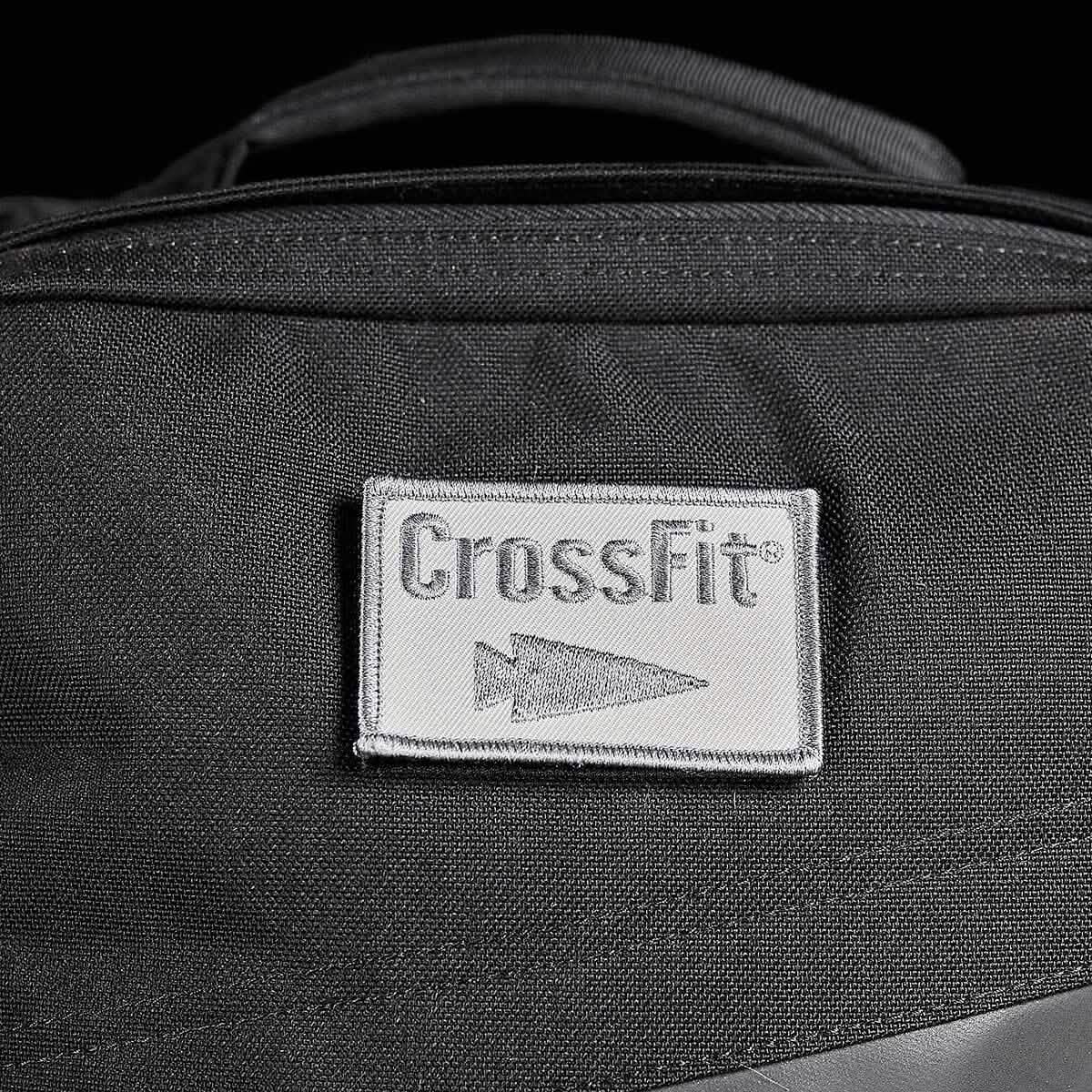 Close-up of a black backpack featuring a gray rectangular Patch - GORUCK x CrossFit from GORUCK-DEMO, showcasing the word "CrossFit" and an arrow logo. The visible fabric texture highlights the stitching details on the embroidered patch and emphasizes its rugged design.