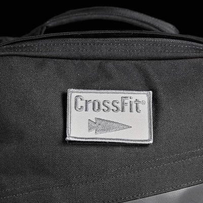 Close-up of a black backpack featuring a gray rectangular Patch - GORUCK x CrossFit from GORUCK-DEMO, showcasing the word "CrossFit" and an arrow logo. The visible fabric texture highlights the stitching details on the embroidered patch and emphasizes its rugged design.