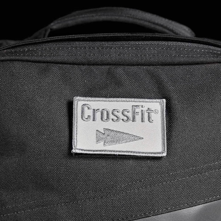 Close-up of a black backpack featuring a gray rectangular Patch - GORUCK x CrossFit from GORUCK-DEMO, showcasing the word "CrossFit" and an arrow logo. The visible fabric texture highlights the stitching details on the embroidered patch and emphasizes its rugged design.