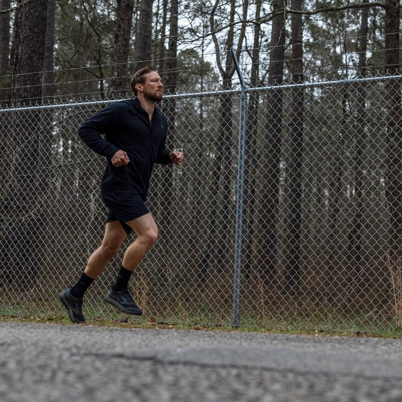 A person is jogging along a paved path near a chain-link fence, surrounded by towering forest trees. They're dressed in a black jacket and shorts, wearing GORUCK's Mackall shoes in Black + Forged Iron, ideal for Special Forces training. The sky is overcast.