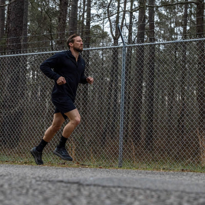 A person is jogging along a paved path near a chain-link fence, surrounded by towering forest trees. They're dressed in a black jacket and shorts, wearing GORUCK's Mackall shoes in Black + Forged Iron, ideal for Special Forces training. The sky is overcast.
