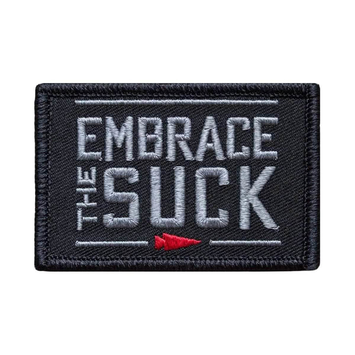 The "Patch - Embrace The Suck" by vendor-unknown features a rectangular design with a black background and prominent gray text stating "EMBRACE THE SUCK." A small red arrow pointing to the right is situated below the text. It also includes a convenient VELCRO backing for easy attachment and removal.