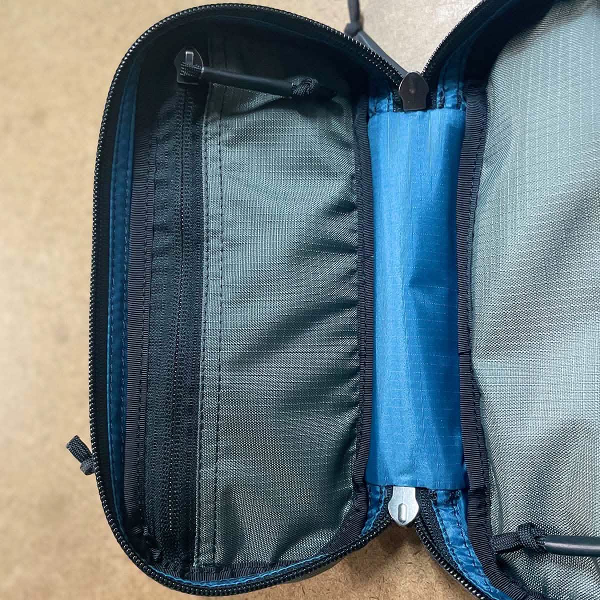 The Bullet Field Pocket Robic Nylon, built by SCARS and crafted by GORUCK, is an open zippered pouch made from robust Robic nylon with a gray interior. It includes various sections such as a mesh pocket on the left and a bright blue band in the center for secure storage. This pouch, recognized for its outstanding tear strength, lies flat on a brown surface.