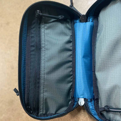 The Bullet Field Pocket Robic Nylon, built by SCARS and crafted by GORUCK, is an open zippered pouch made from robust Robic nylon with a gray interior. It includes various sections such as a mesh pocket on the left and a bright blue band in the center for secure storage. This pouch, recognized for its outstanding tear strength, lies flat on a brown surface.
