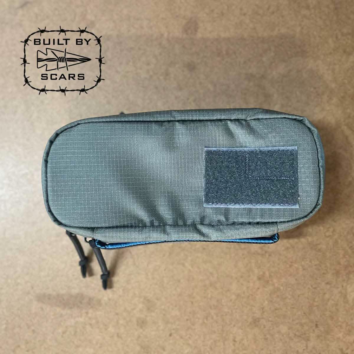 A GORUCK Bullet Field Pocket, crafted from robust Robic nylon in green, lies on a textured surface. The top of the pocket is secured with Velcro and features a logo of barbed wire with an arrow, along with the words "BUILT BY SCARS," emphasizing its exceptional tear strength.