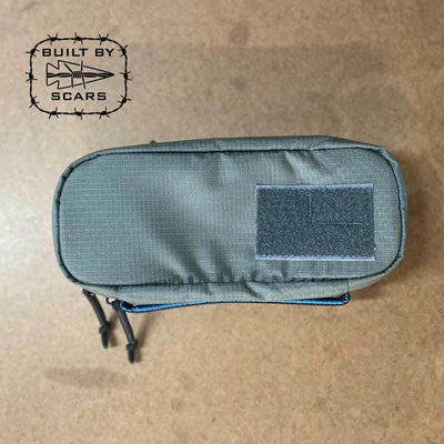 A GORUCK Bullet Field Pocket, crafted from robust Robic nylon in green, lies on a textured surface. The top of the pocket is secured with Velcro and features a logo of barbed wire with an arrow, along with the words "BUILT BY SCARS," emphasizing its exceptional tear strength.