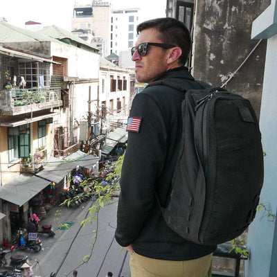 A man wearing sunglasses stands with his GR1 USA - Cordura (The Original Ruck) backpack from GORUCK on a balcony, recalling the intensity of his last GORUCK Challenge as he surveys the bustling street below.