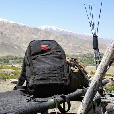 A black GORUCK GR1 USA - Cordura (The Original Ruck) sits on a bench alongside archery arrows, its bombproof laptop compartment primed for any adventure, all framed by a breathtaking mountainous backdrop.