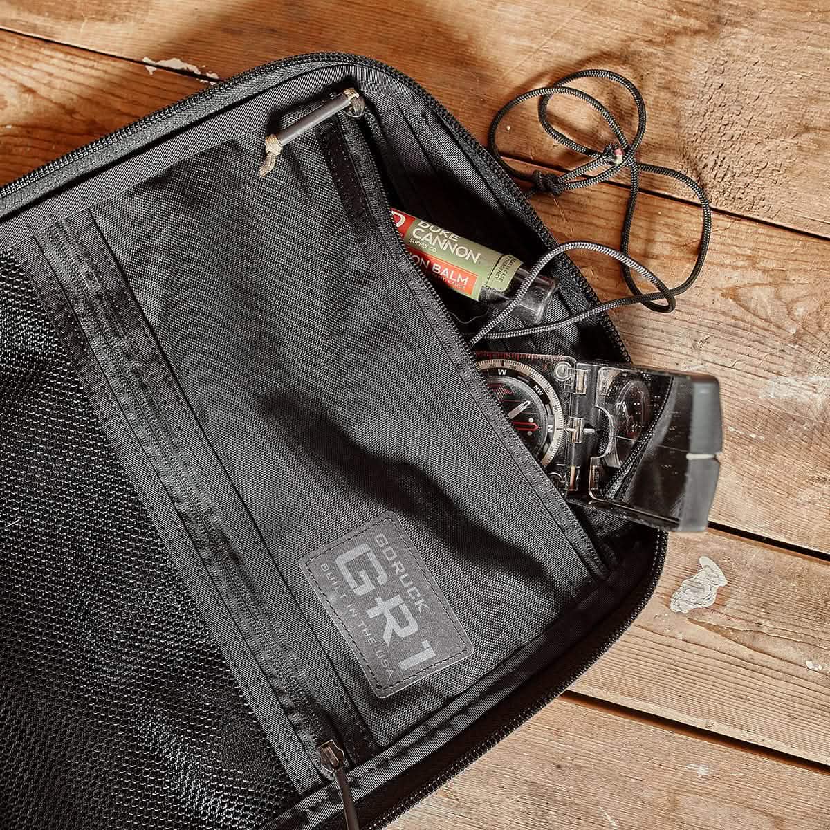A GR1 USA - Cordura from GORUCK sits open on the wooden surface, revealing a compass and lip balm—essentials for any Special Forces mission, epitomizing the quality of American manufacturing.