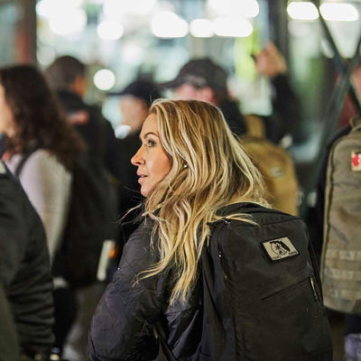 A woman with long blonde hair and a GR1 USA rucksack by GORUCK, known for its Special Forces-grade durability, stands in a crowded area, looking to the side.