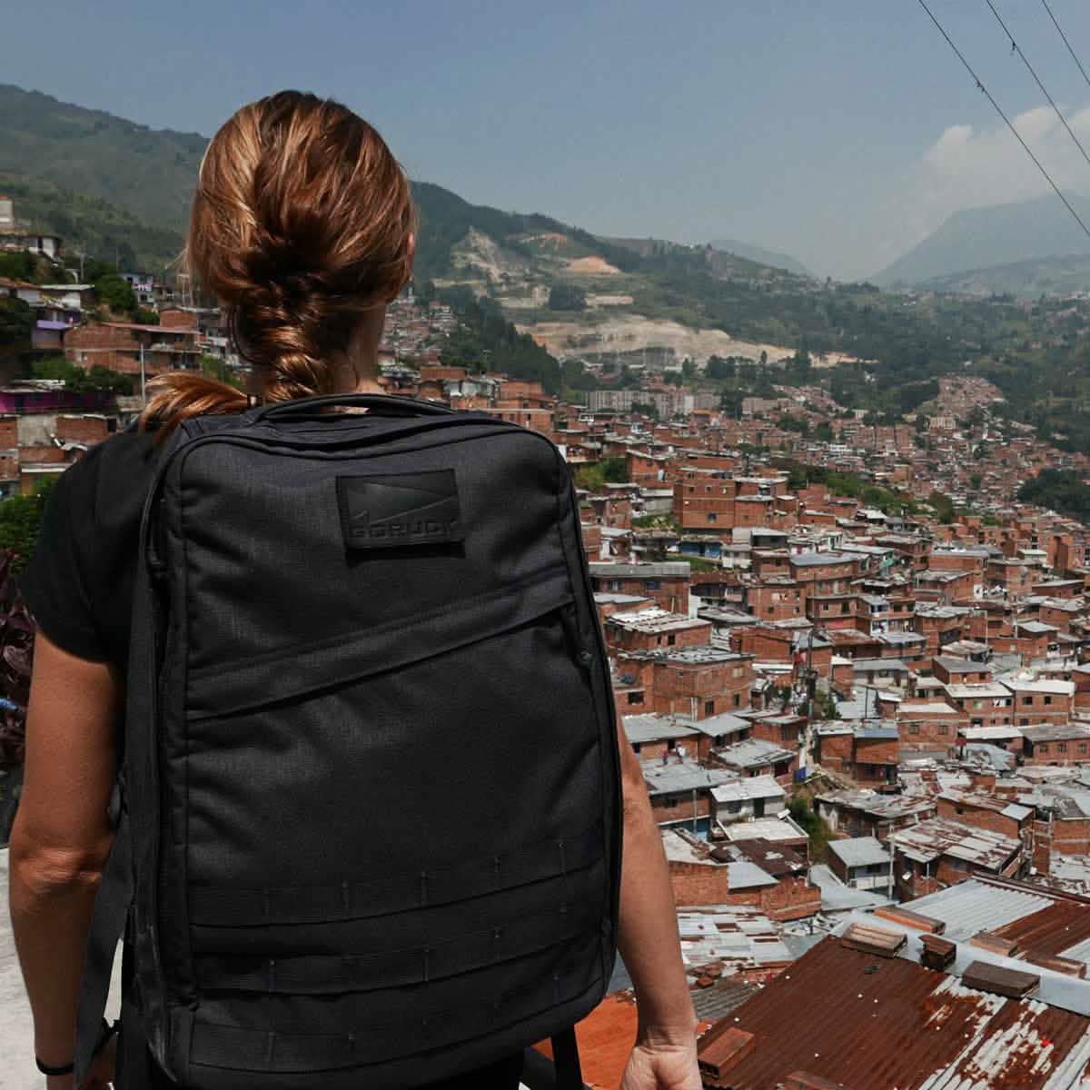 A person carrying the GR1 USA rucksack by GORUCK gazes over a densely packed hillside cityscape under a clear sky.