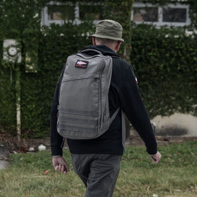 A person sporting a hat and a GR1 USA - Cordura rucksack by GORUCK strolls by a vine-covered building, the bag's bombproof laptop compartment ensuring everything inside remains safe and secure.