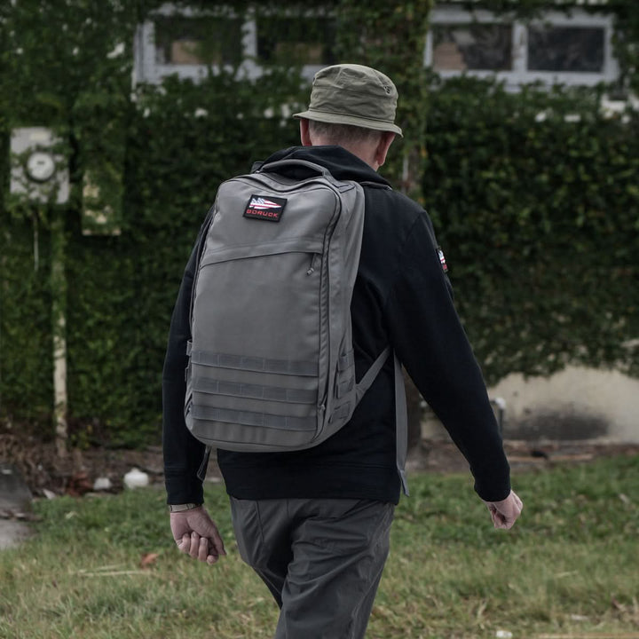 A person sporting a hat and a GR1 USA - Cordura rucksack by GORUCK strolls by a vine-covered building, the bag's bombproof laptop compartment ensuring everything inside remains safe and secure.