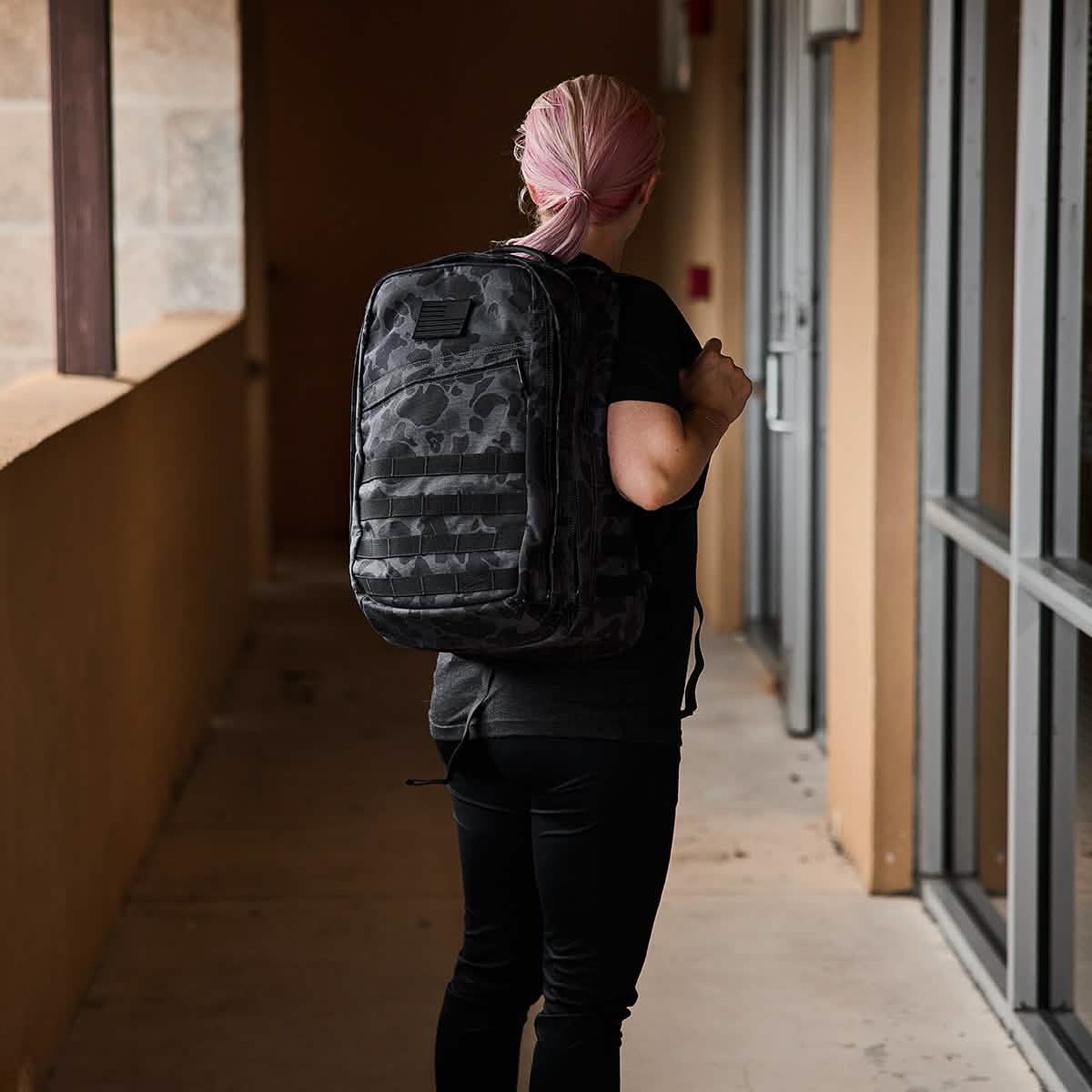 A person with pink hair is walking down a hallway adorned with beige walls and windows on one side, wearing a black GORUCK GR2 featuring a camouflage pattern reminiscent of Special Forces gear. They are dressed in a black shirt and pants, with their back turned toward the camera.