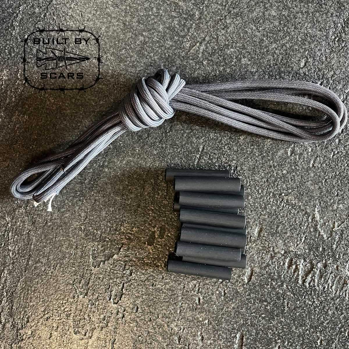 A Paracord Kit - Built By SCARS from GORUCK, featuring a coiled gray paracord and several small black heat shrink tubes ideal for DIY ruck upgrades, is neatly arranged on a textured gray surface with "Built by SCARS" printed in the top left corner alongside the logo.