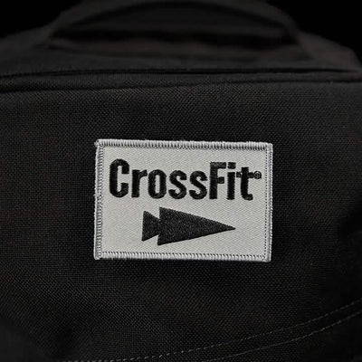 A close-up of a black fabric displays the Patch - GORUCK x CrossFit from GORUCK-DEMO, featuring an embroidered white square with the word "CrossFit" in bold black letters. Below it, a sleek GORUCK arrowhead symbol points to the right, signifying strength and movement.