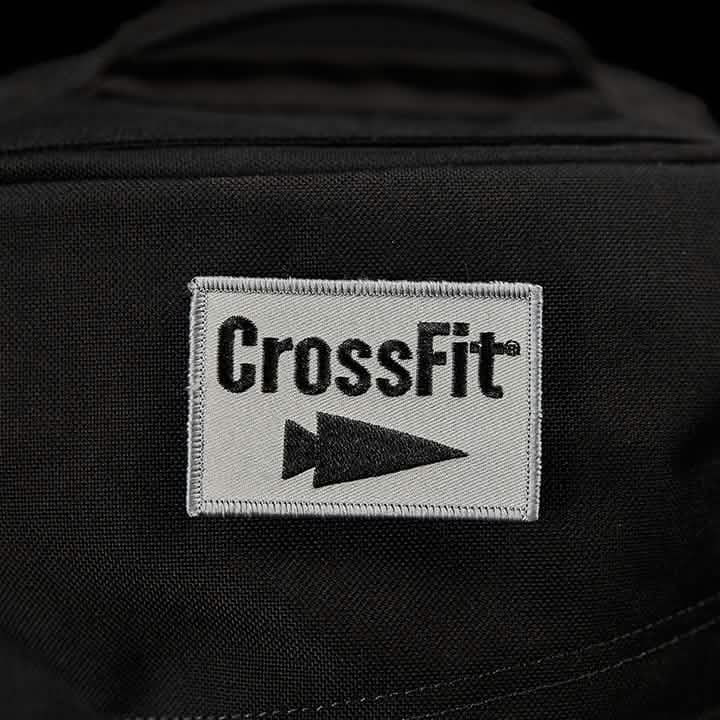 A close-up of a black fabric displays the Patch - GORUCK x CrossFit from GORUCK-DEMO, featuring an embroidered white square with the word "CrossFit" in bold black letters. Below it, a sleek GORUCK arrowhead symbol points to the right, signifying strength and movement.