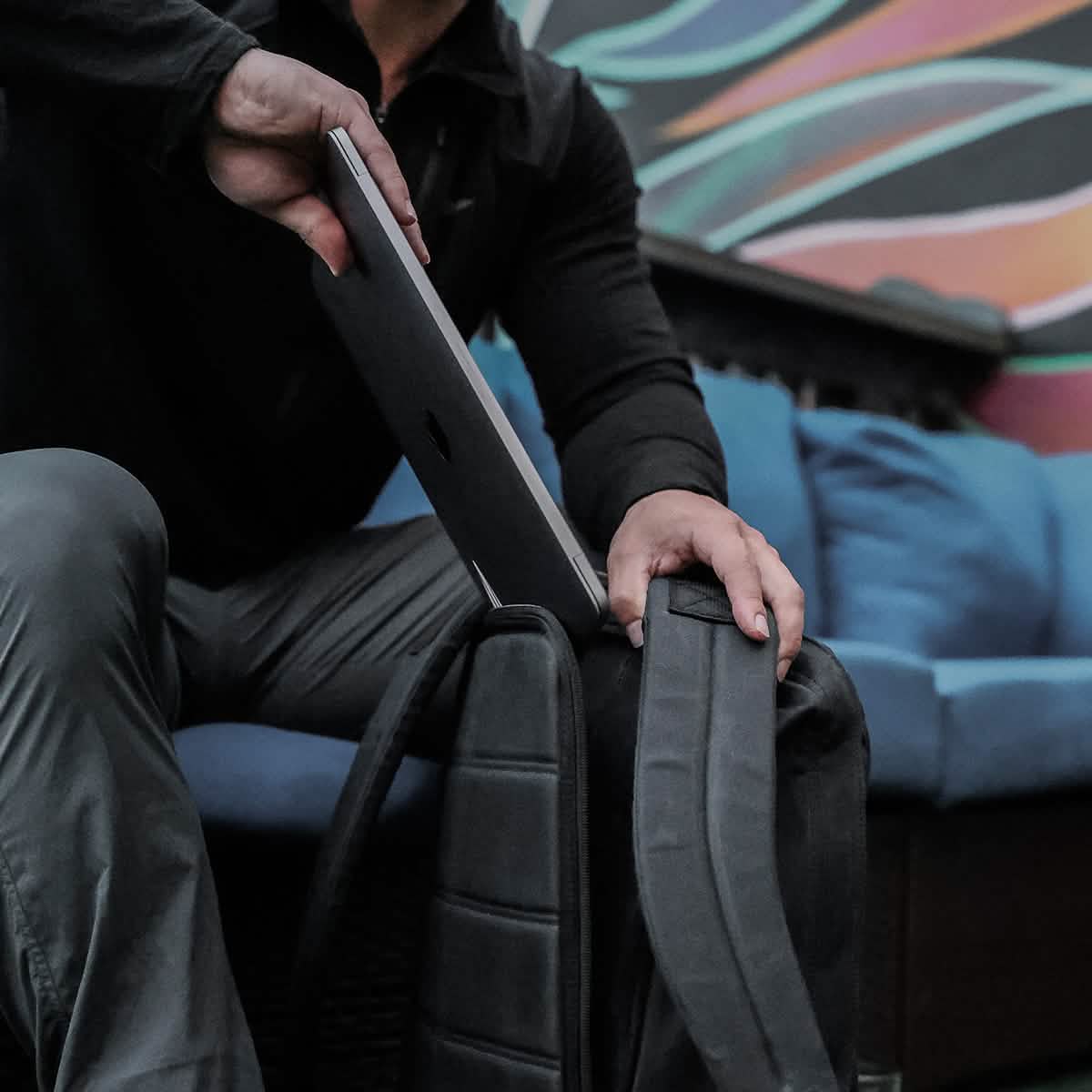 A person places a closed laptop into a GR1 USA - Heritage rucksack by GORUCK while sitting indoors. The background features a colorful mural and a blue cushioned sofa. The individual is wearing a black long-sleeve shirt and gray pants.