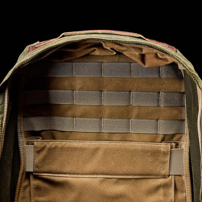 The image showcases a GORUCK GR1 USA - Heritage Backpack with an open view of its interior. The inside is brown, featuring horizontal straps and a large pocket for organizing belongings. The exterior is crafted from olive green fabric, echoing a retro canvas rucksack style with visible zippers.