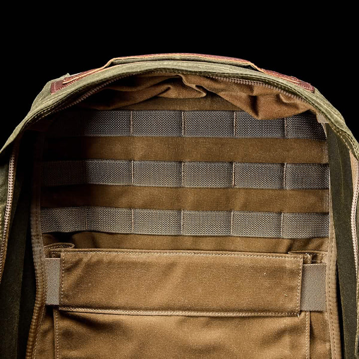 The image showcases a GORUCK GR1 USA - Heritage Backpack with an open view of its interior. The inside is brown, featuring horizontal straps and a large pocket for organizing belongings. The exterior is crafted from olive green fabric, echoing a retro canvas rucksack style with visible zippers.