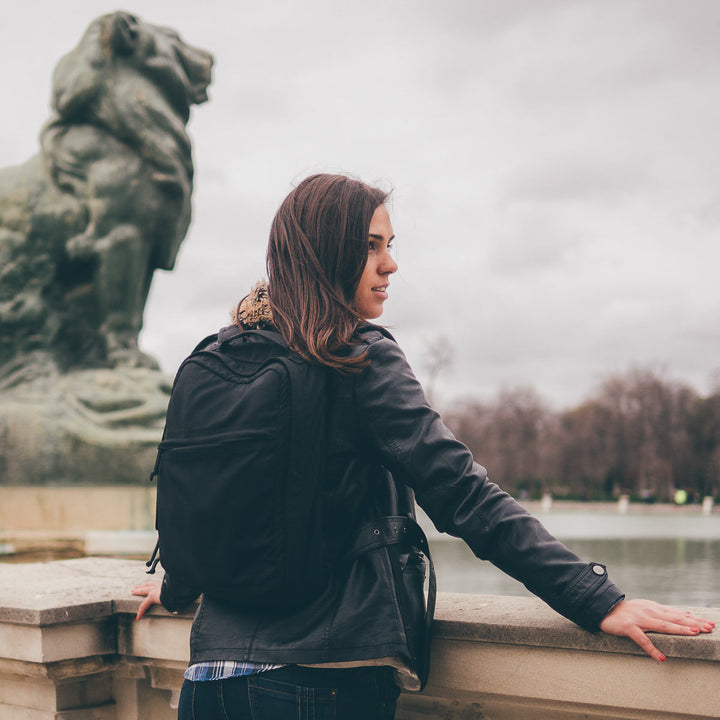 Woman with a GR0 USA Slick - Ballistic Nylon Cordura backpack, featuring a bombproof laptop compartment, leans on a stone railing near a lion statue and pond on a cloudy day.
