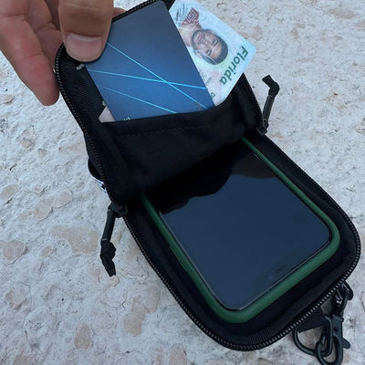 A hand reveals a black GORUCK Phone Pouch featuring a zippered closure, showcasing a smartphone, a partially visible credit card, and a photo ID. The scene is set outdoors on a stone-like surface.