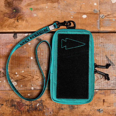 A Phone Pouch by GORUCK in teal fabric, crafted from durable CORDURA, features a black Velcro front panel and zipper closures. It rests on a rustic, paint-stained wooden table. The pouch includes an adjustable teal strap with metal clasps for convenience.