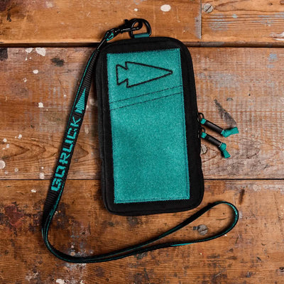 A Phone Pouch from GORUCK, featuring a teal and black color combination and arrow design, rests on a rustic wooden surface. It includes a matching teal detachable lanyard adorned with the GORUCK logo in bold letters. Crafted from 500D CORDURA, the pouch boasts a rugged, outdoor appearance.