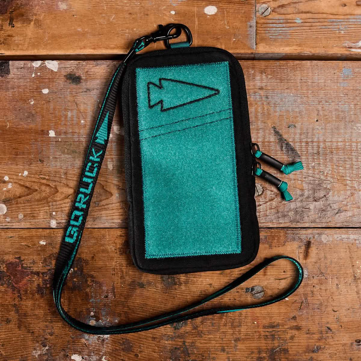 A Phone Pouch from GORUCK, featuring a teal and black color combination and arrow design, rests on a rustic wooden surface. It includes a matching teal detachable lanyard adorned with the GORUCK logo in bold letters. Crafted from 500D CORDURA, the pouch boasts a rugged, outdoor appearance.