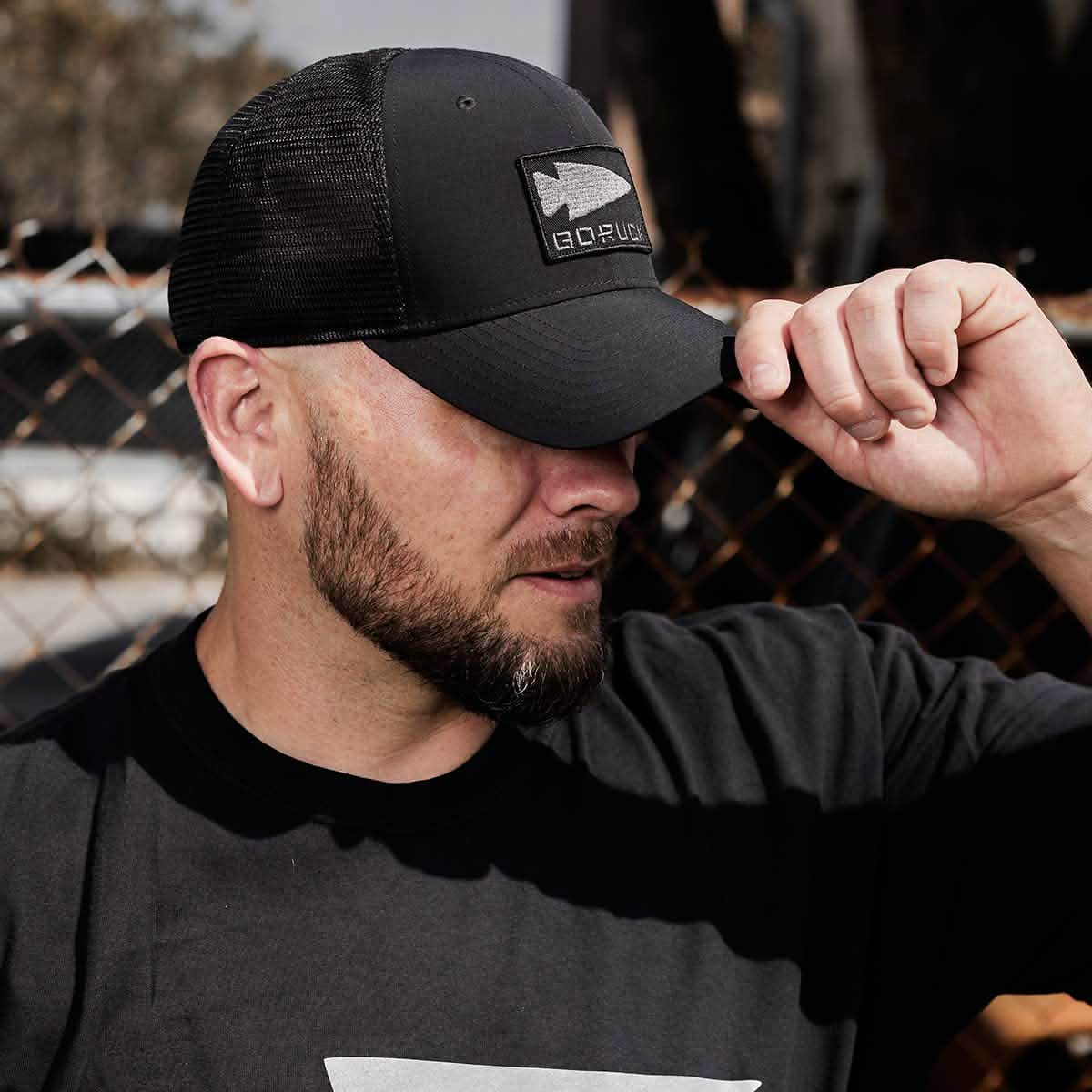 A bearded man in a black T-shirt and mesh trucker hat stands outdoors. He adjusts the brim of his GORUCK Performance Trucker Hat, adorned with a Spearhead patch, as a chain-link fence is visible in the background.
